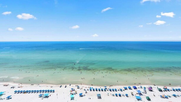 A beach with clear turquoise water, a sandy shore, and numerous blue umbrellas and lounge chairs. - Majestic T2-1606 - Fabulous Three Bedrooms Condo! (Panama City Beach)