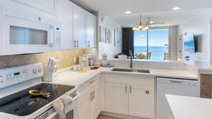 A modern kitchen with white cabinetry, a stove, microwave, and a dishwasher. There is a large window offering a view of the ocean. - Majestic T2-1606 - Fabulous Three Bedrooms Condo! (Panama City Beach)