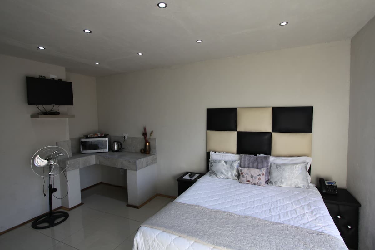 A modern hotel room with a large bed, a flat-screen TV mounted on the wall, a small kitchenette, and a bedside table with a phone.