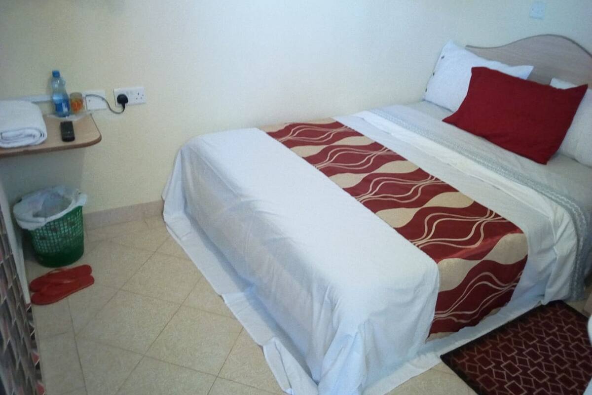 Una cama individual con una colcha blanca y roja estampada, un almoháraco rojo y un cabecero de madera. Hay una mesita con una toalla doblada y una botella de agua, una cesta verde y un objeto rojo en el suelo.