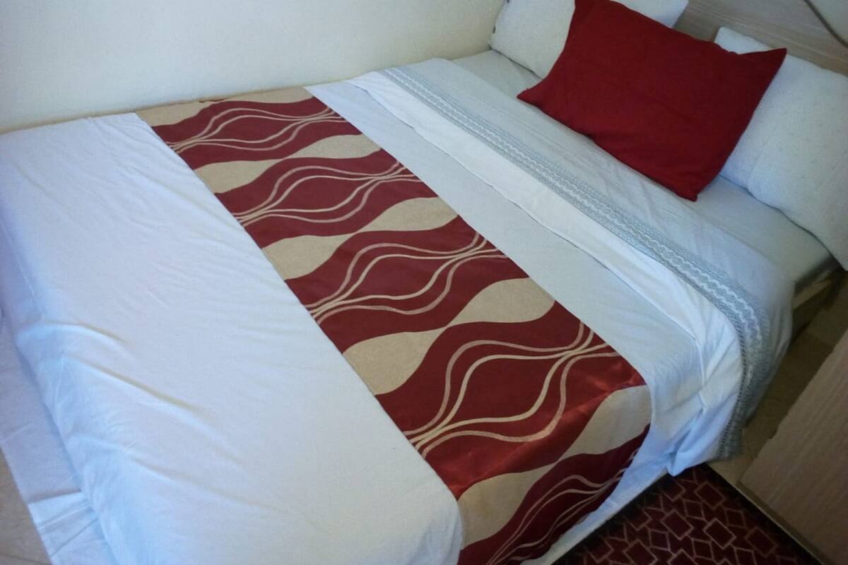 Una cama individual con una colcha estampada, almohadas blancas y un cojín decorativo rojo.