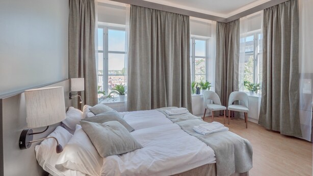 A modern hotel room with a large bed, two chairs, a window with curtains, and a view of the city. - Hamnhotellet (Vastervik)