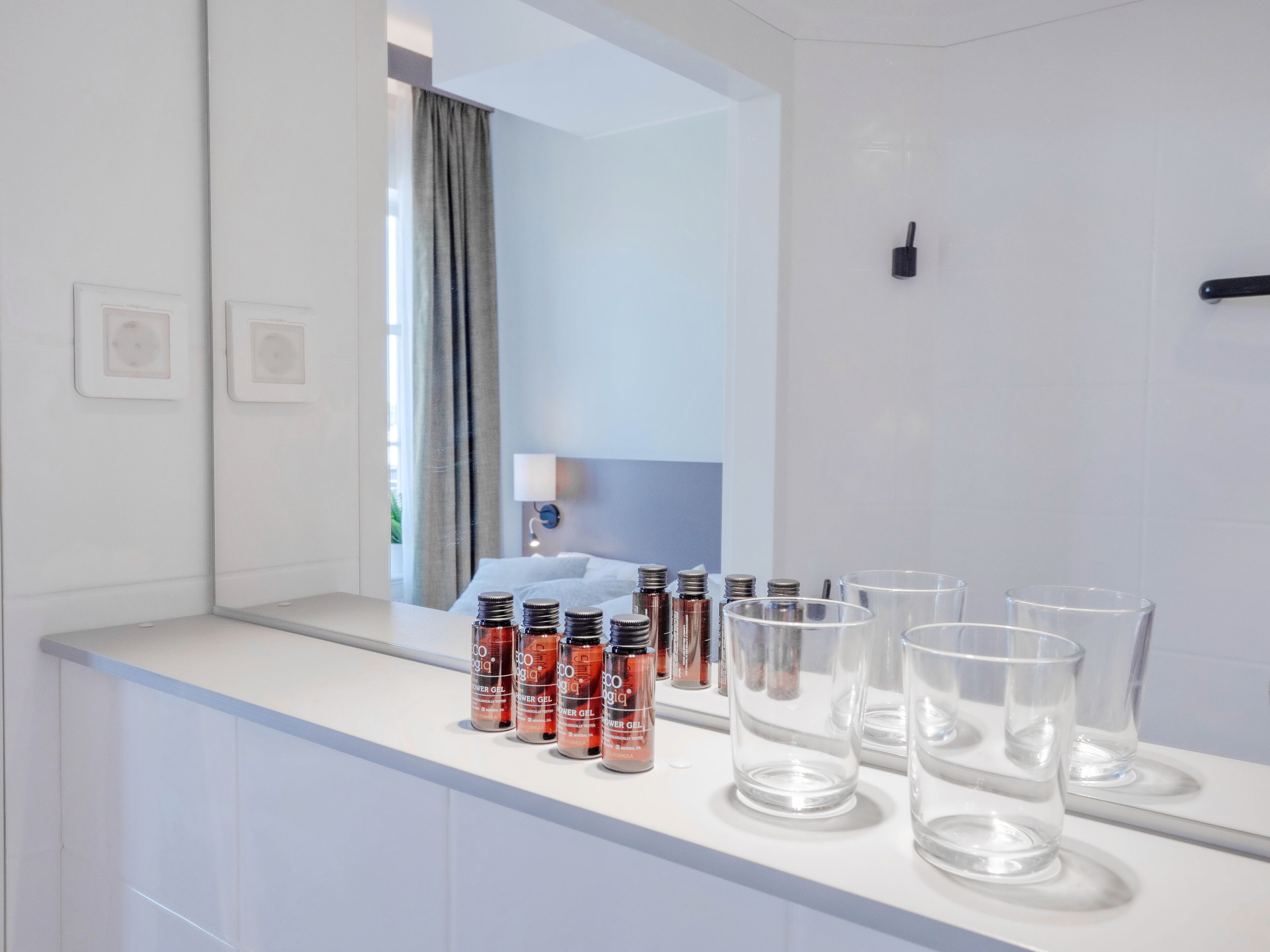 Un baño con un mostrador blanco, un espejo y una ventana con cortinas. Hay varias botellas y vasos sobre el mostrador.