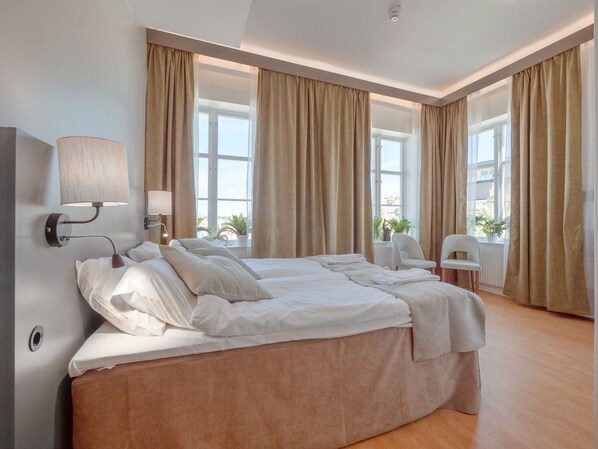 A hotel room with a large bed, two bedside tables with lamps, a chair, and large windows with curtains. - Hamnhotellet (Vastervik)