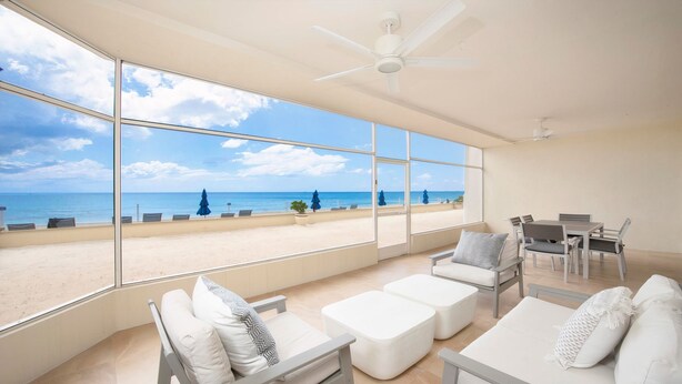 A spacious room with large windows overlooking a beach, furnished with white sofas, a dining table, and a ceiling fan. - Laguna Del Mar (Seven Mile Beach)
