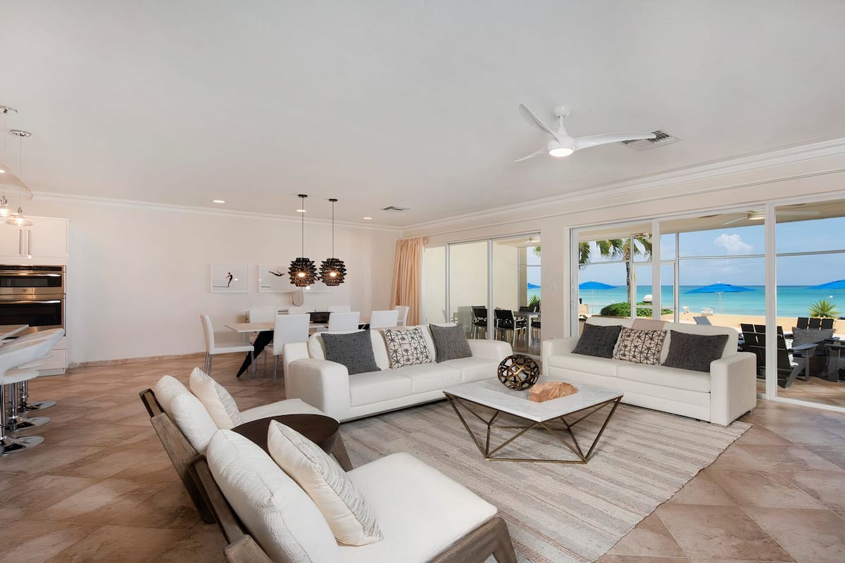Ein modernes Wohnzimmer mit einer weißen Sofagarnitur, einem Couchtisch und einem Essbereich mit Tisch und Stühlen. Ein großes Fenster bietet Blick auf den Strand und den Ozean.