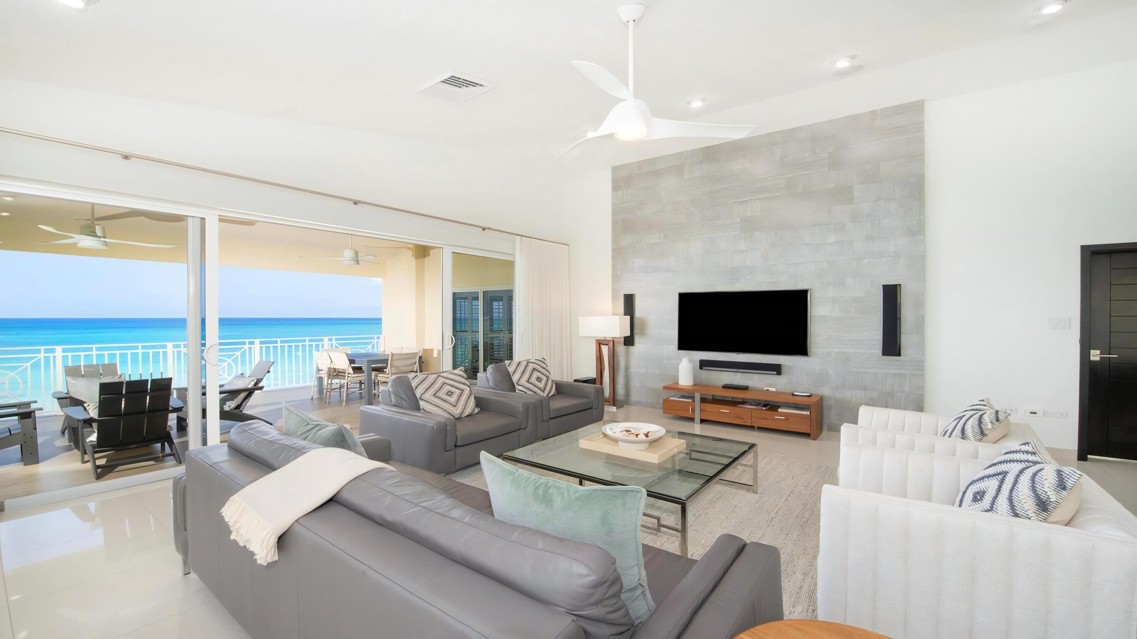 Ein modernes Wohnzimmer mit einem großen Flachbildfernseher, grauen Sofas, einem Couchtisch und einem Balkon mit Blick auf das Meer.