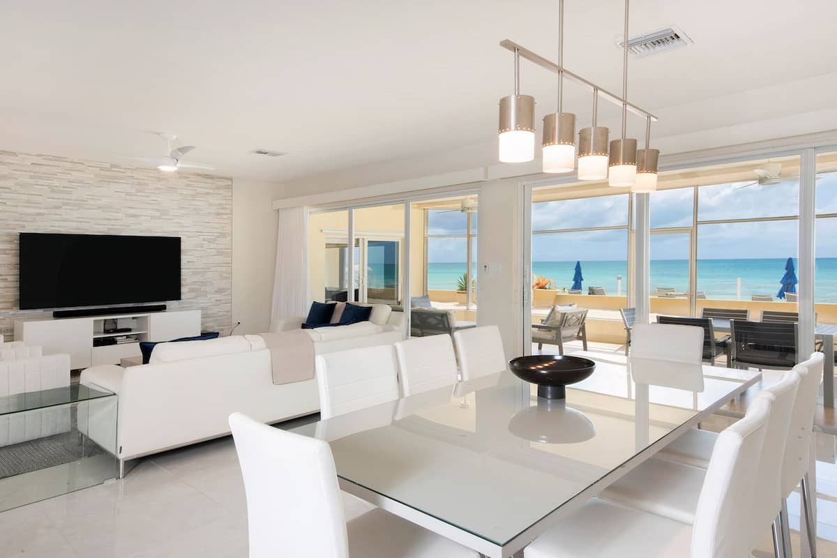 Ein modernes Wohnzimmer mit Fernseher, weißen Sofas, einem Glastisch und großen Fenstern mit Blick auf den Strand.