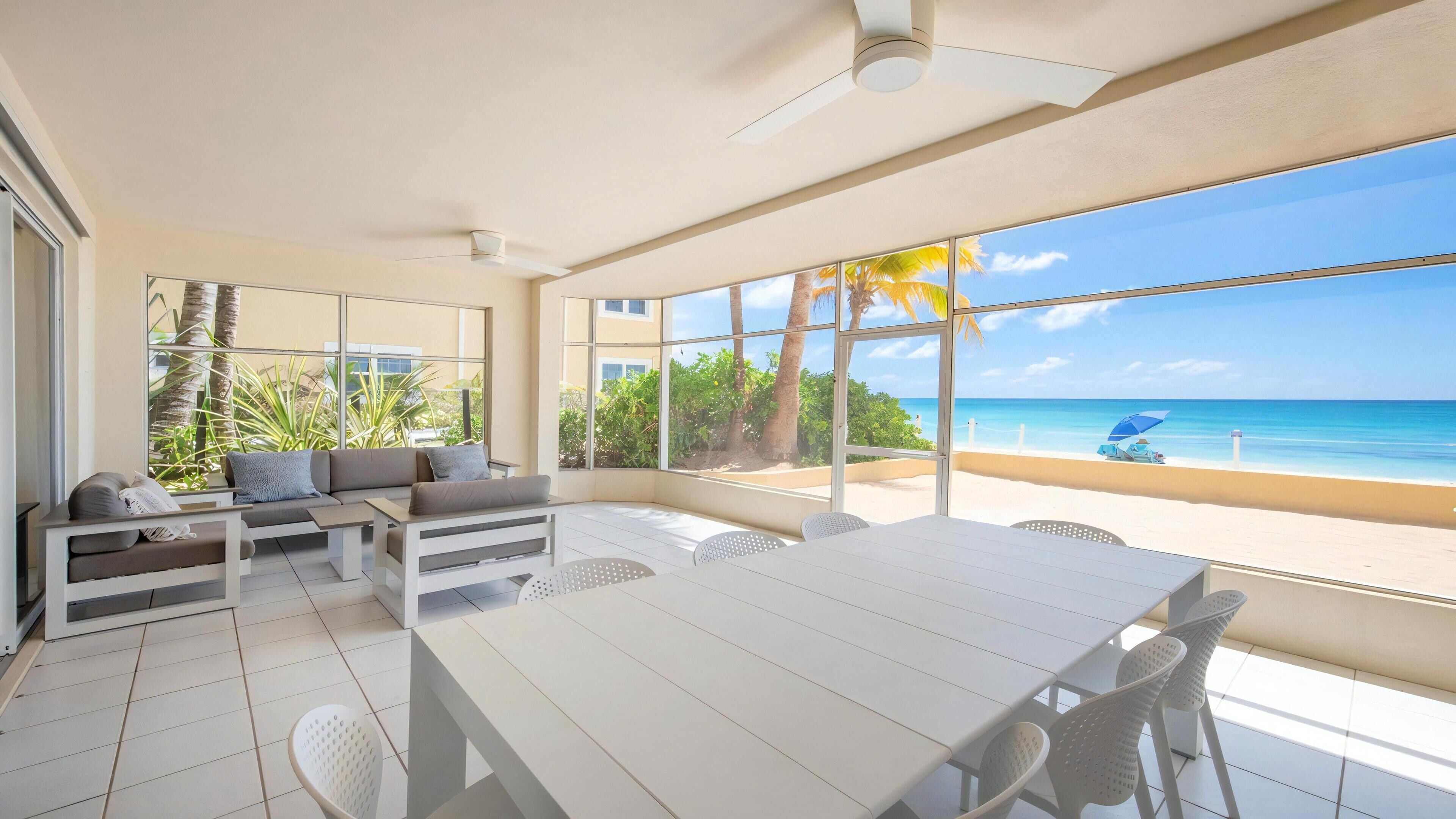 A spacious room with a large window overlooking a beach, a white dining table, and a seating area with a sofa and chairs.