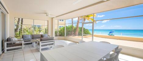 A spacious room with a large window overlooking a beach, a white dining table, and a seating area with a sofa and chairs.