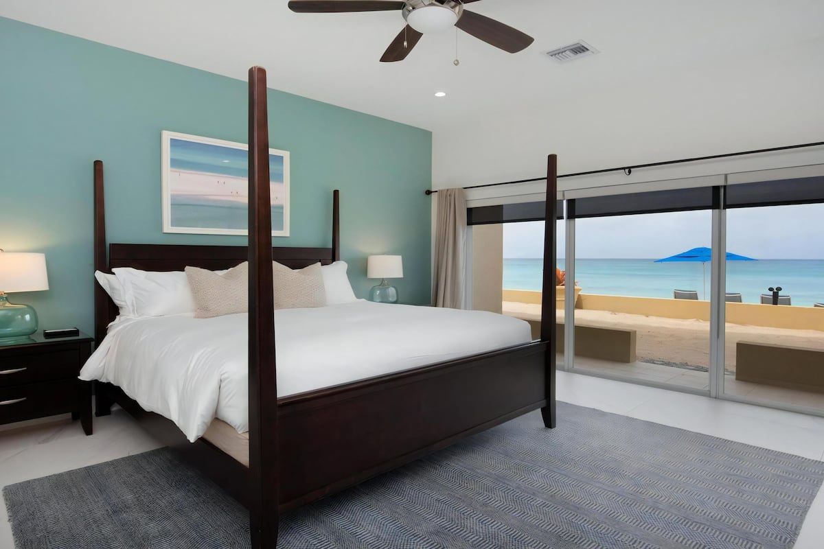 Ein Schlafzimmer mit Himmelbett, Nachttisch mit Lampe, Beistelltisch mit Lampe und großem Fenster mit Blick auf Strand und Meer.