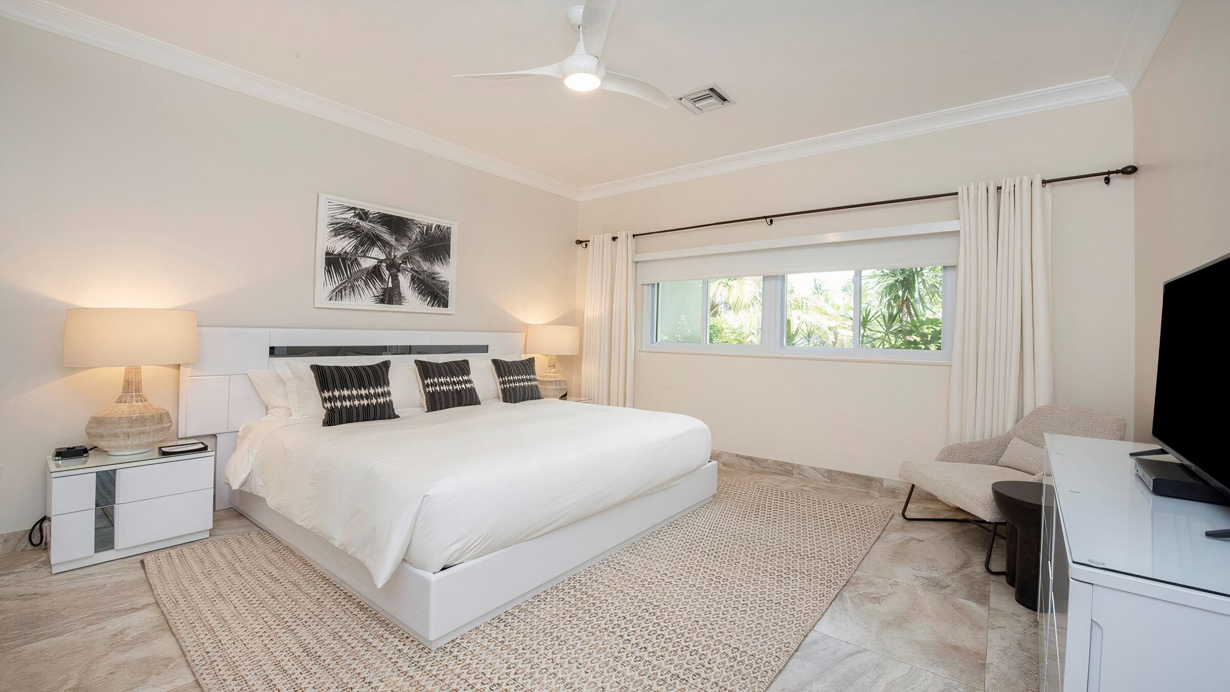 Une chambre moderne avec un grand lit, des tables de chevet, un ventilateur de plafond, une fenêtre avec des rideaux et un téléviseur.
