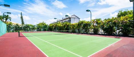 En tennisbane med net, omgivet af grønt og en bygning i baggrunden.