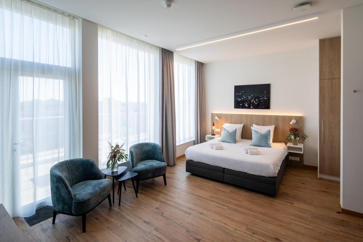 A modern hotel room with a large bed, two armchairs, a small table, and a nightstand.