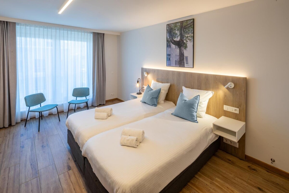 A hotel room with two beds, a desk, a chair, and a large window with curtains.