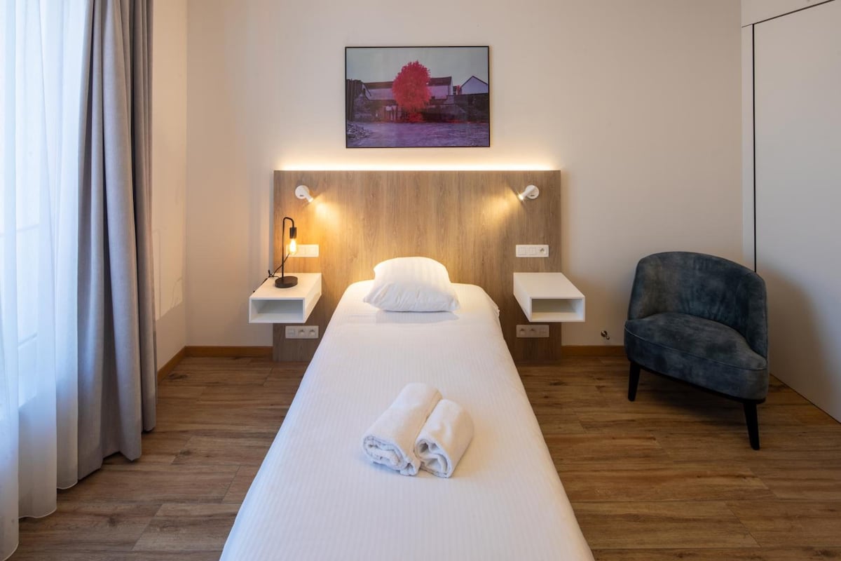 A hotel room with a single bed, a bedside table with a lamp, a chair, and a painting on the wall.