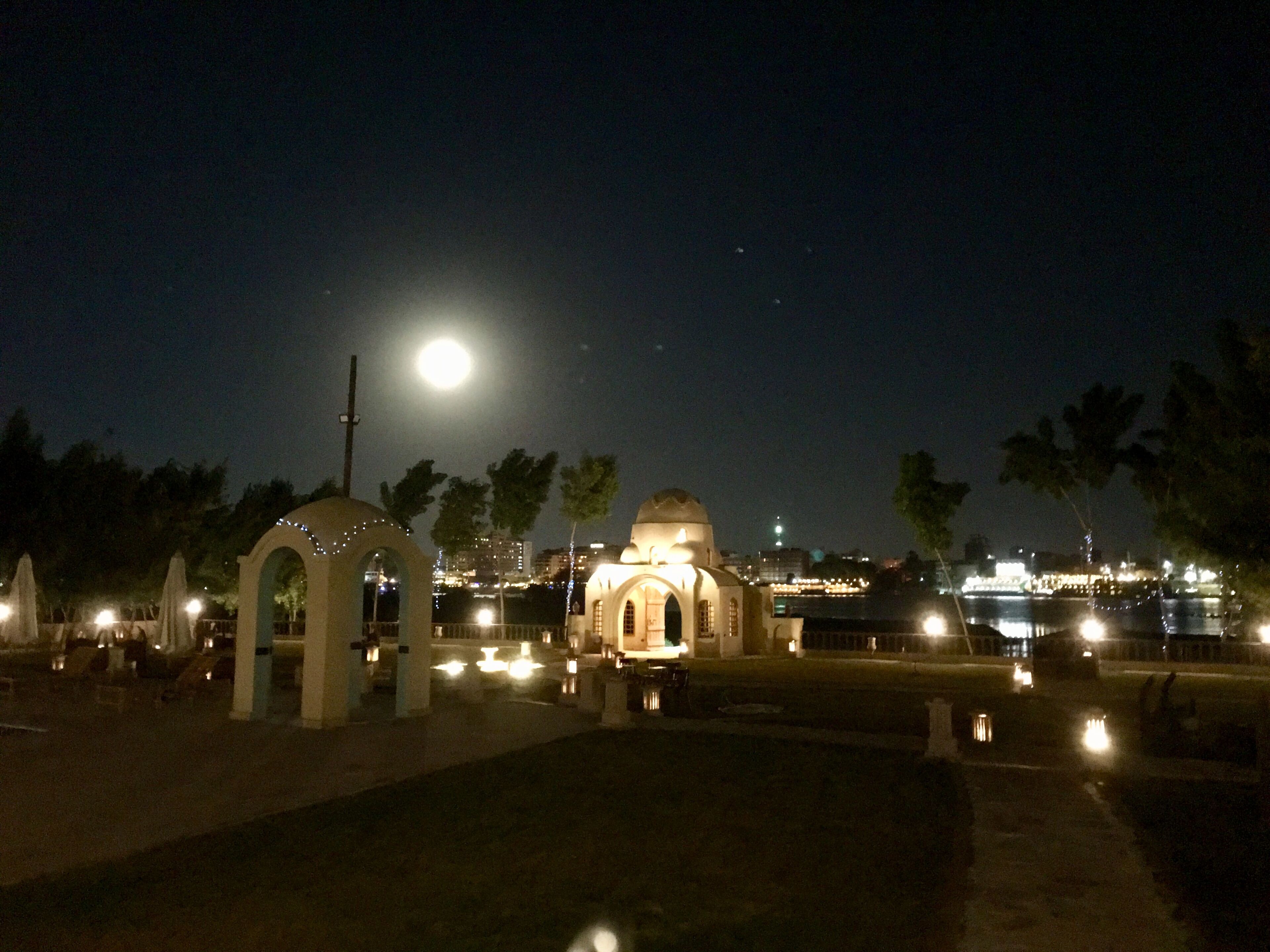 Une scène nocturne avec un bâtiment illuminé, une lune et une étendue d’eau.
