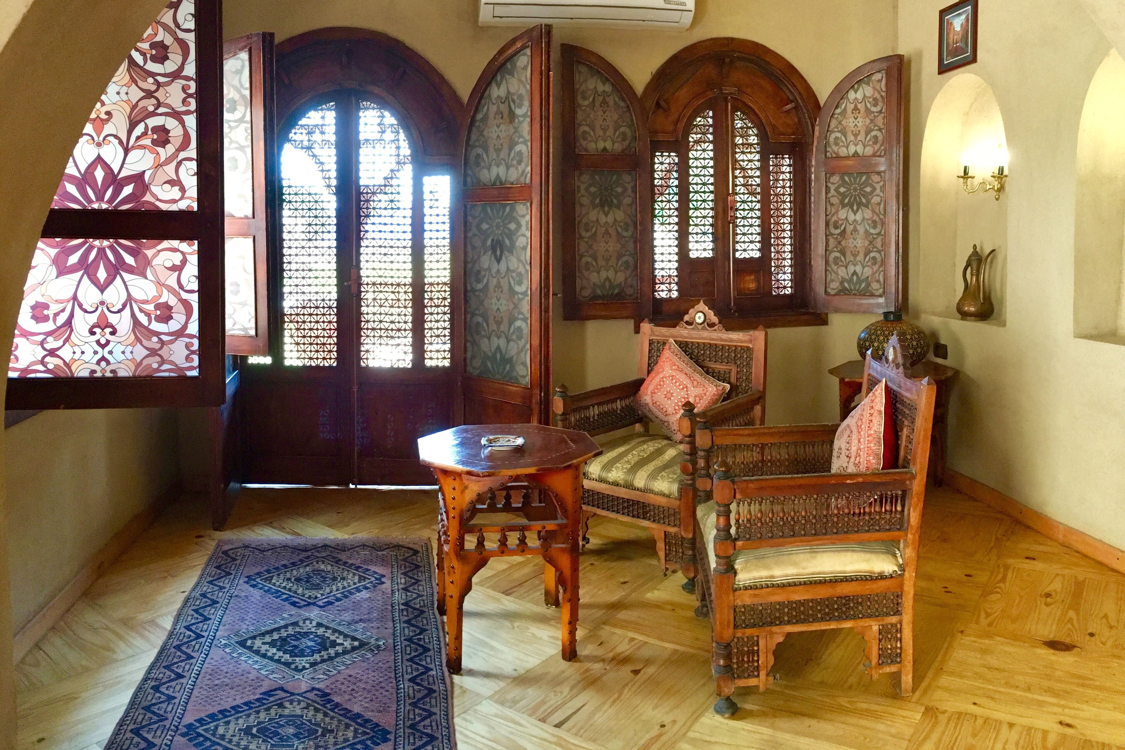 Una habitación tradicional con suelo de madera, un tapiz estampado, una silla de madera, una mesita pequeña y una pared decorativa con ventanales de arco.