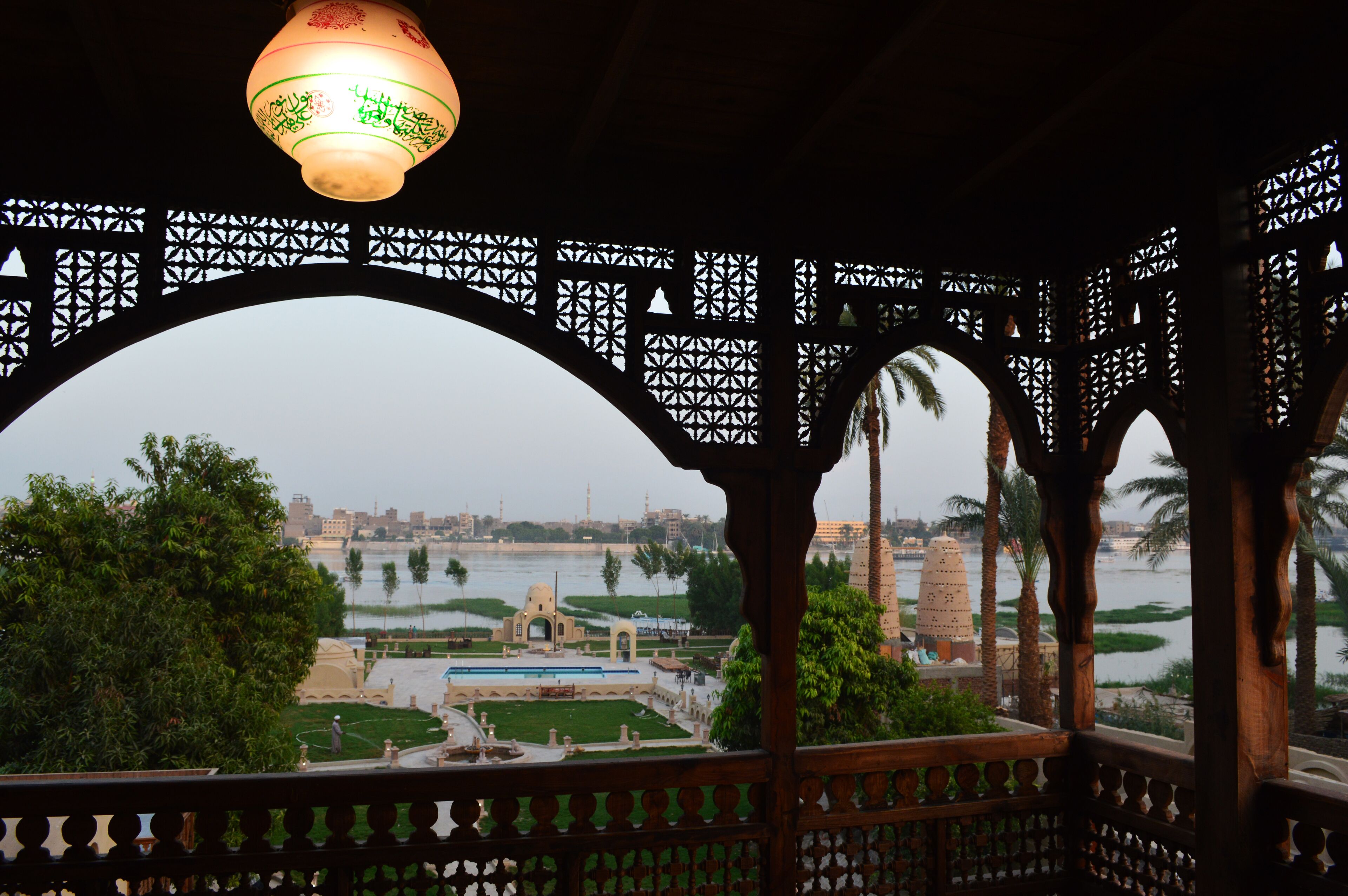 A view from a balcony with a decorative wooden railing, overlooking a river, palm trees, and a distant cityscape.