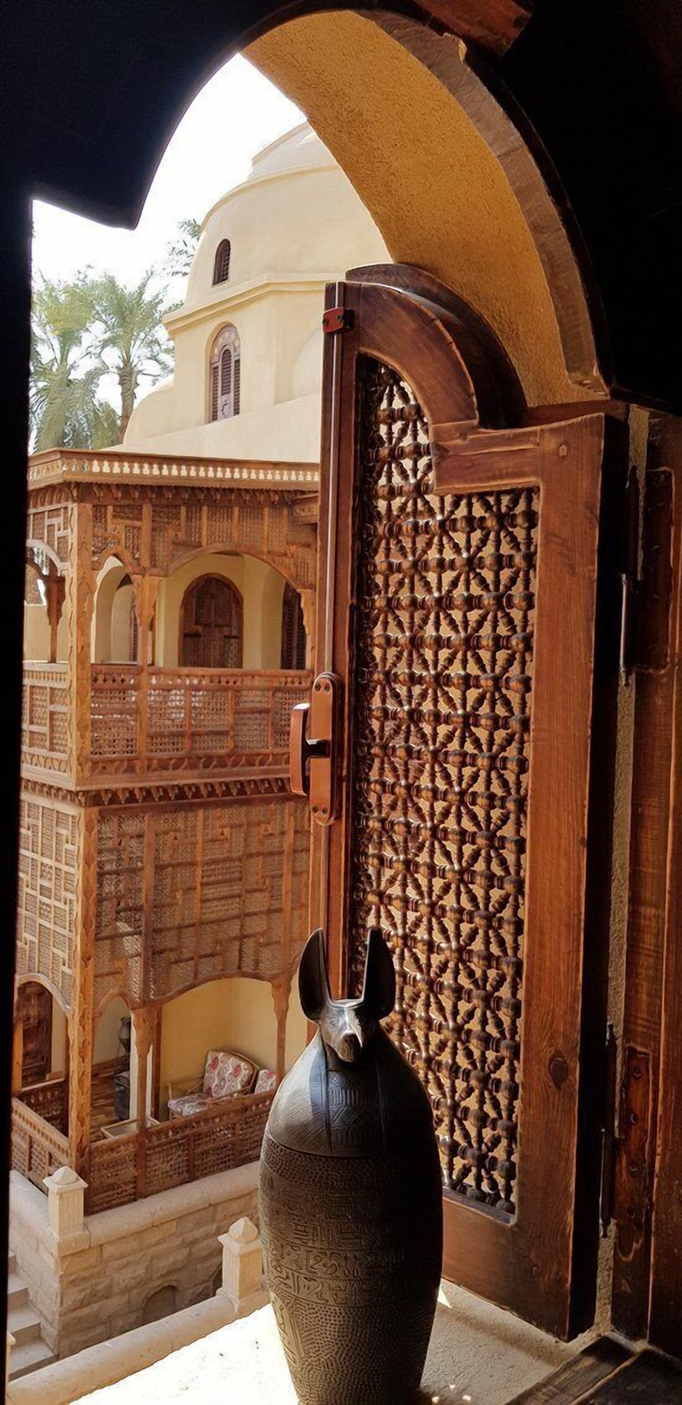 A traditional wooden door with intricate carvings, leading to a courtyard with a white building and a large vase.