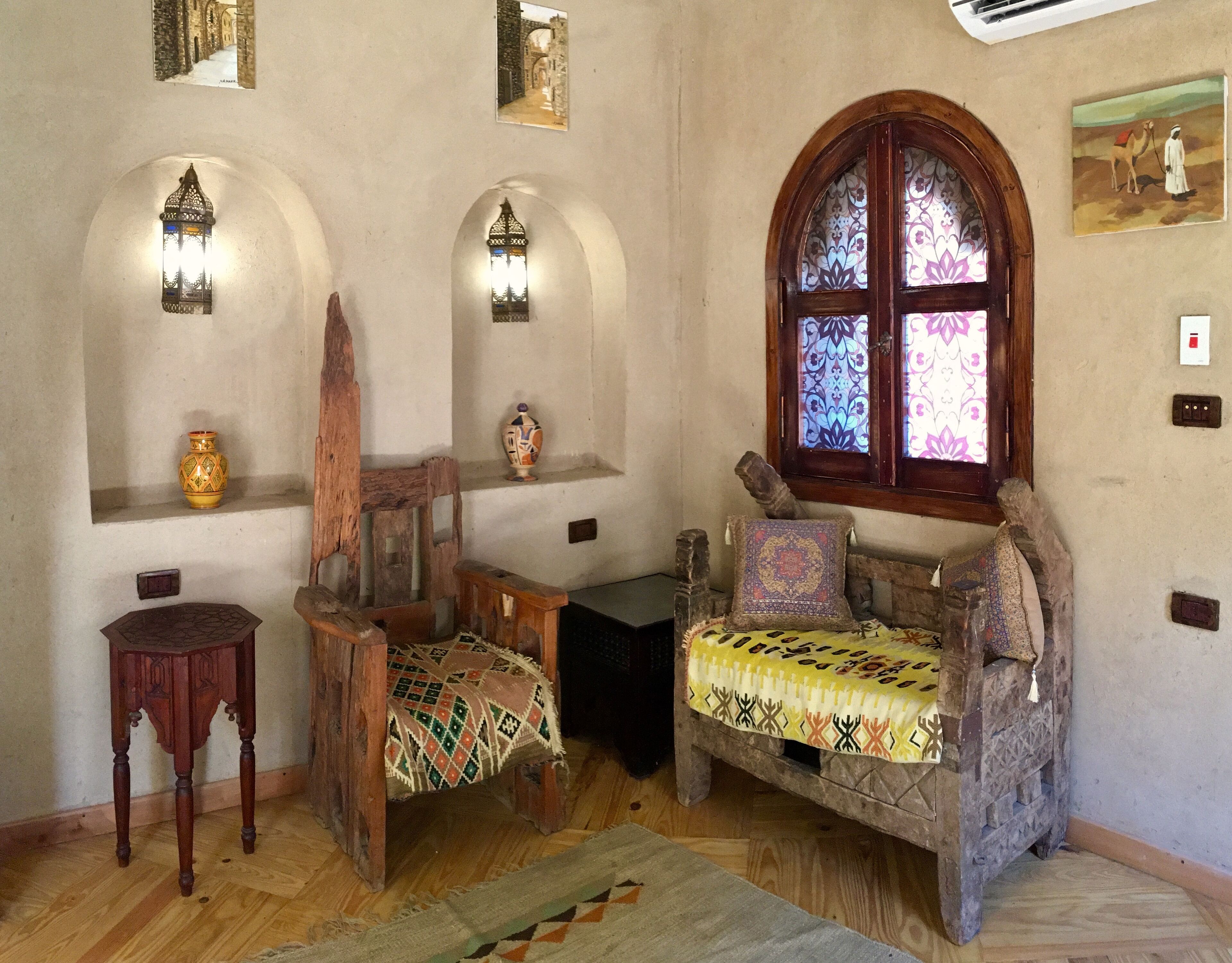 A room with a wooden chair, a small table, a bench with a patterned cushion, and a large arched window with stained glass.