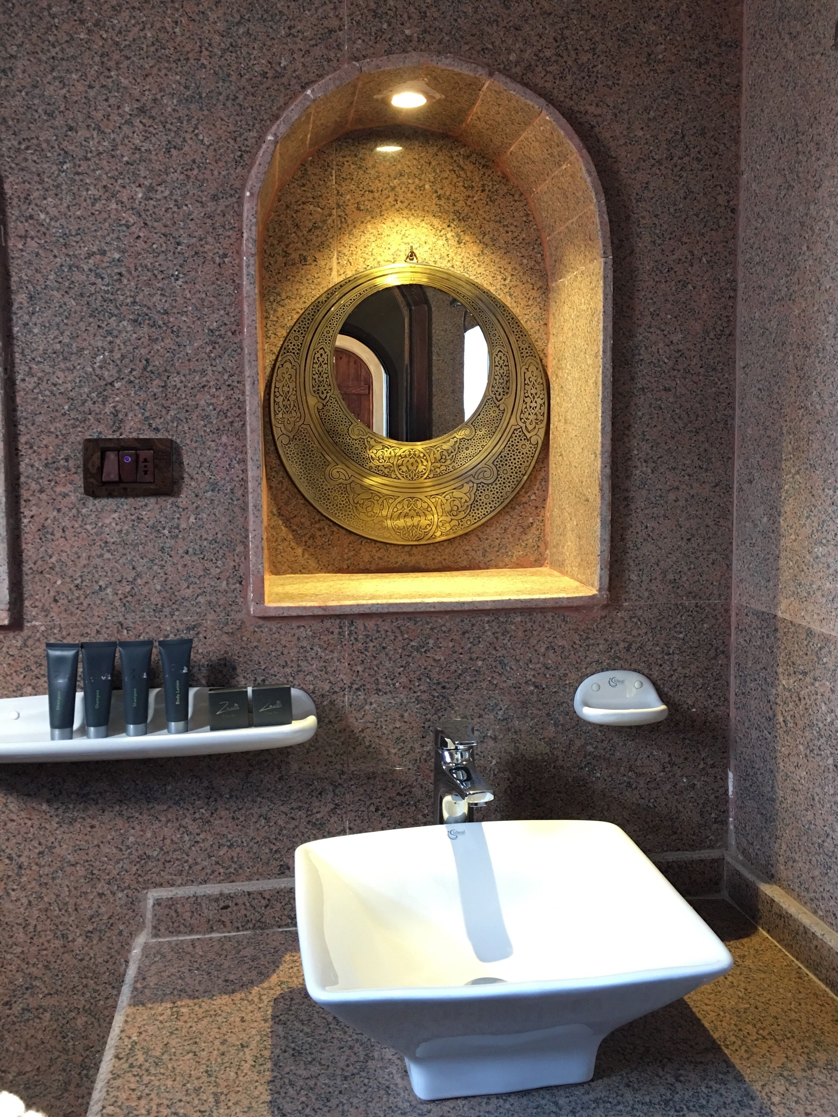 Ein Badezimmer mit einem weißen Waschbecken, einem runden Spiegel mit aufwendigen Verzierungen und einer kleinen Ablage mit Pflegeprodukten.