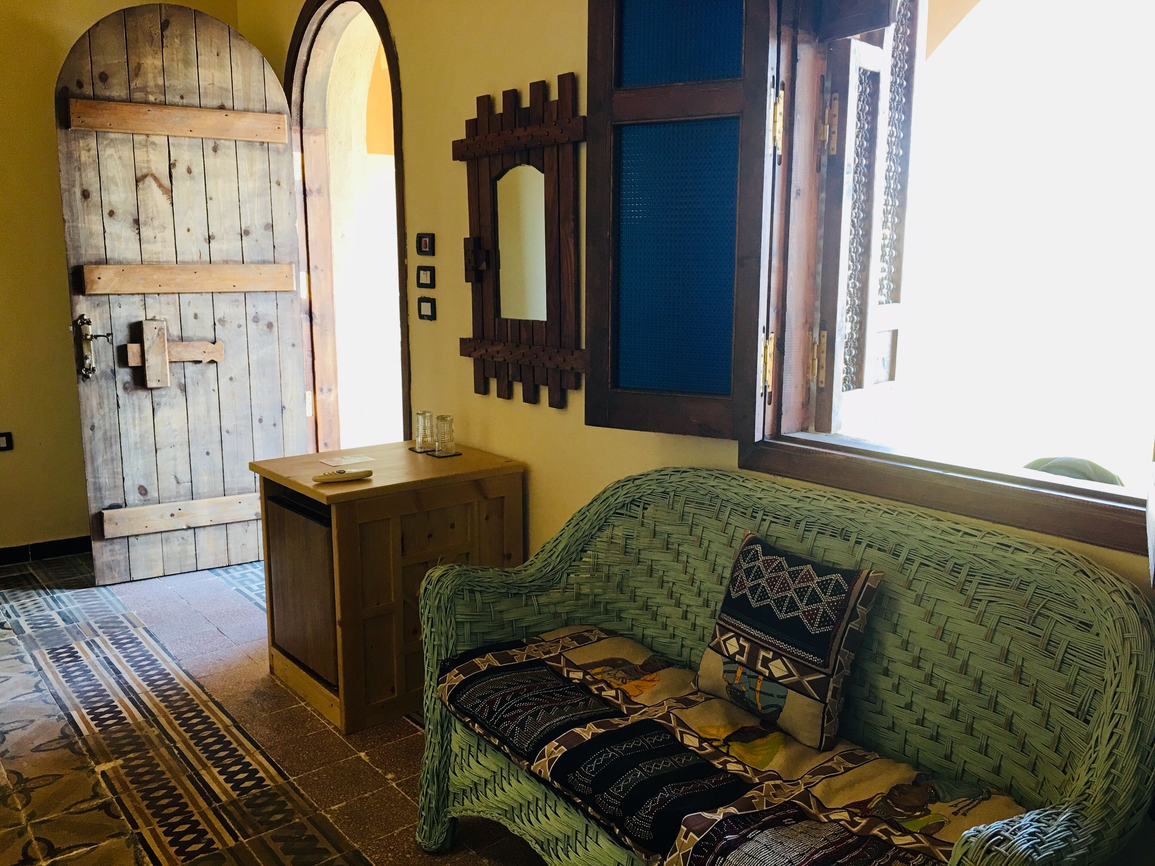 Um quarto com porta de madeira, um sofá de vime com almofadas estampadas, uma pequena mesa de madeira e um espelho na parede.