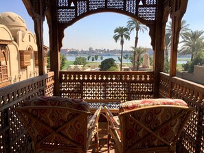 A balcony with two patterned chairs, a view of a cityscape, palm trees, and a body of water. - Djorff Palace (Luxor)