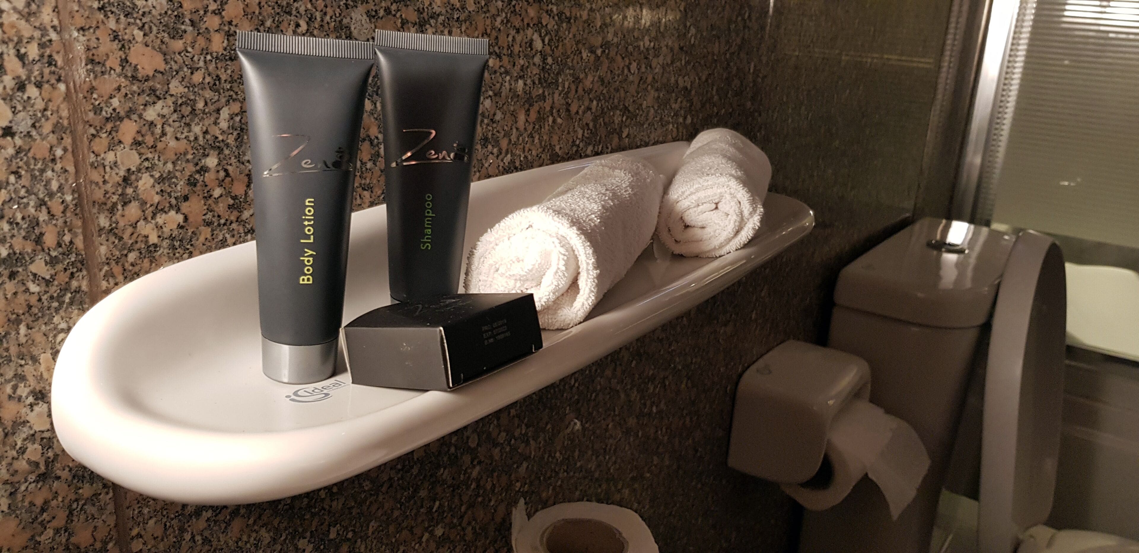 Un estante de baño con artículos de aseo, como loción corporal, champú y una caja, junto con toallas dobladas.