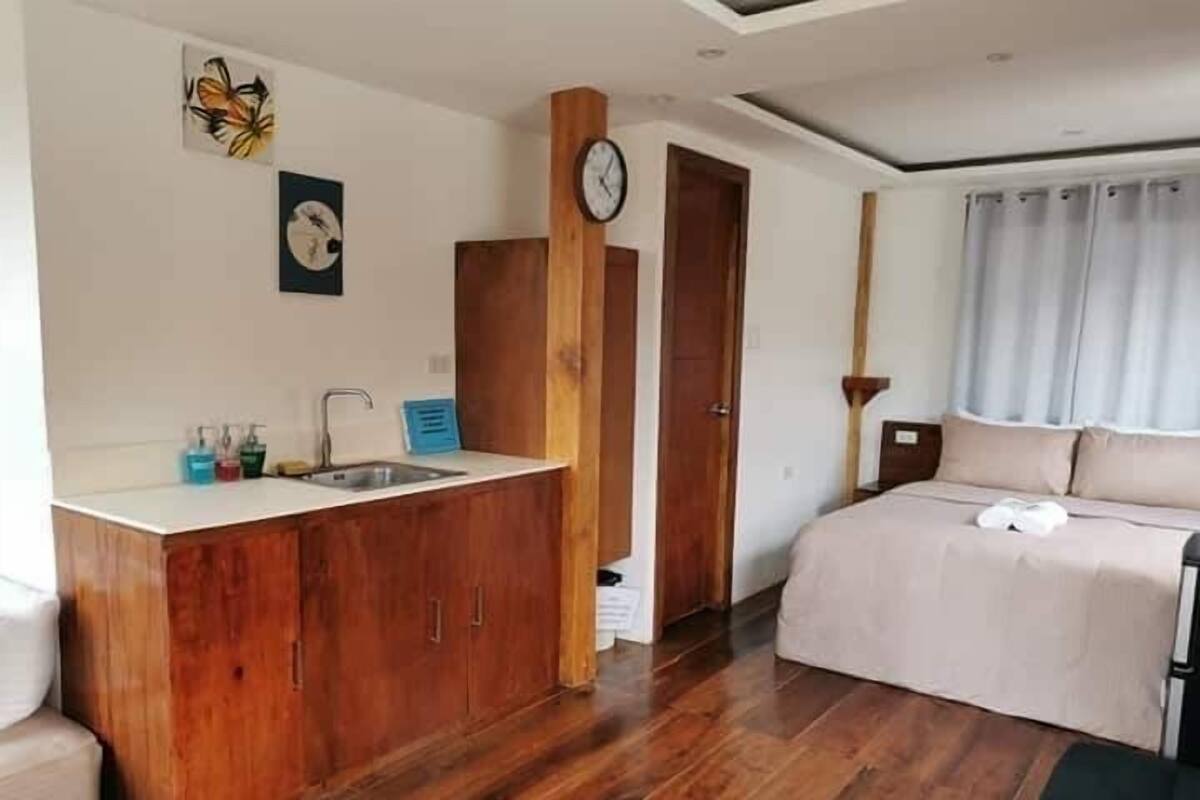A bedroom with a wooden wardrobe, a bed, a nightstand, a clock, and a window with curtains.