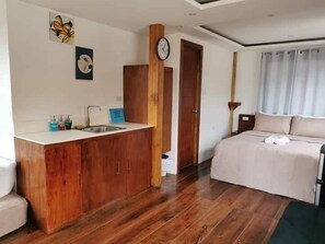 A bedroom with a wooden wardrobe, a bed, a nightstand, a clock, and a window with curtains.