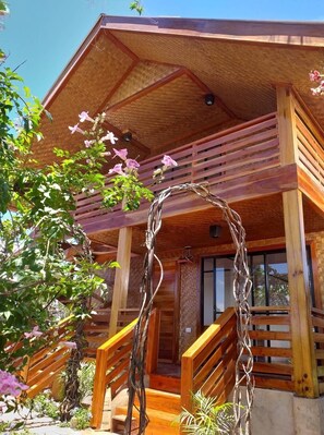 A wooden cabin with a balcony, wooden stairs, and a garden area with flowers. - Descanso en Paraiso Resort (San Juan)