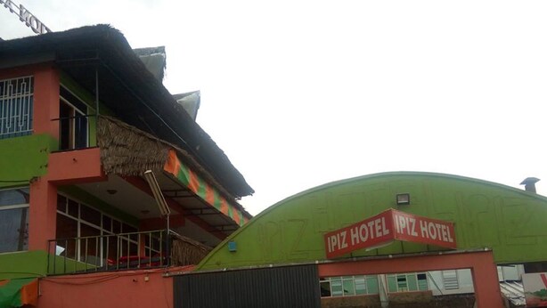 Een hotel met een geel-oranje gevel, een bord waarop 'IPIZ HOTEL' staat, en terraszitplaatsen met parasols.