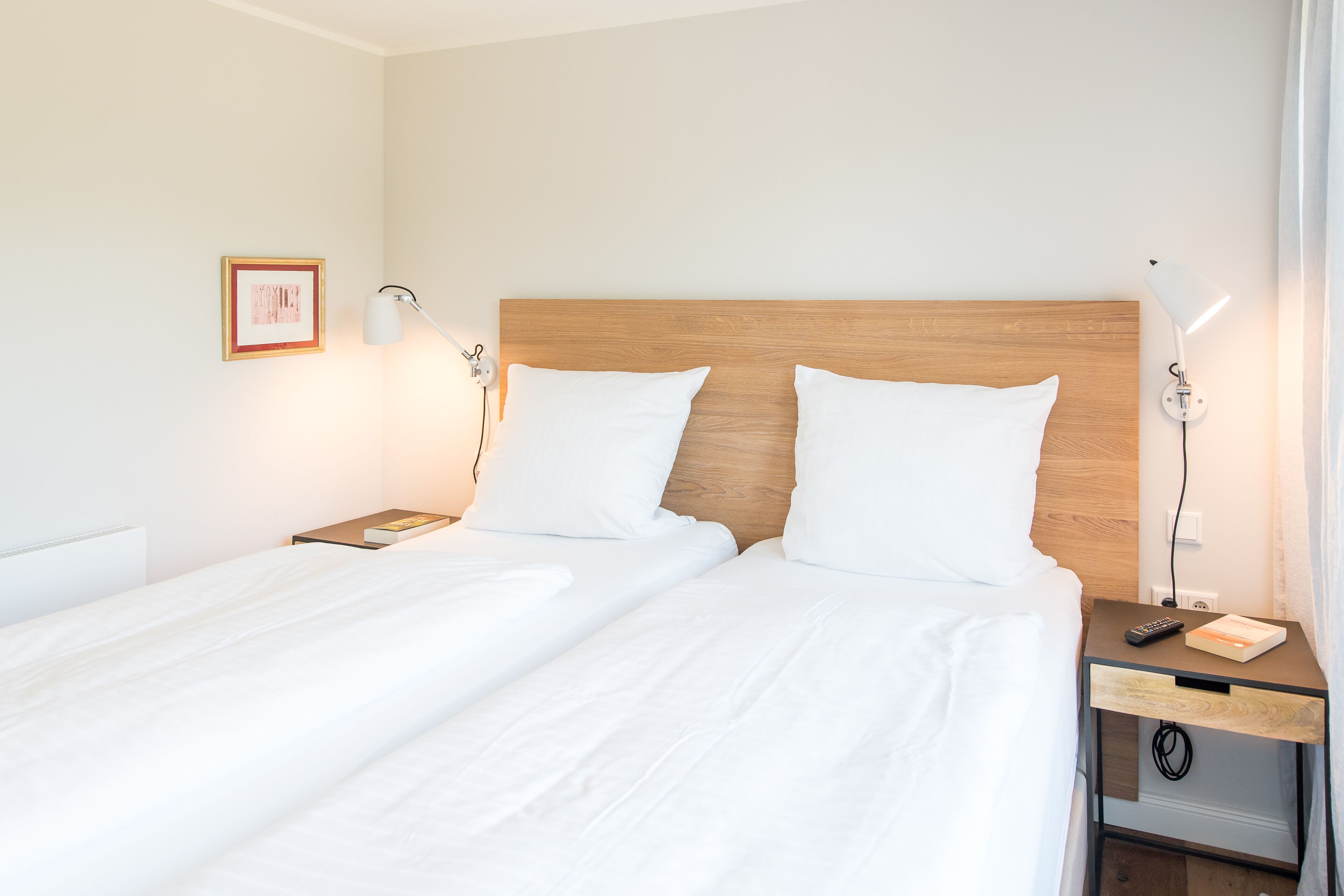A double bed with white linens, two bedside tables with lamps, a framed picture on the wall, and a small nightstand with a book and remote control.