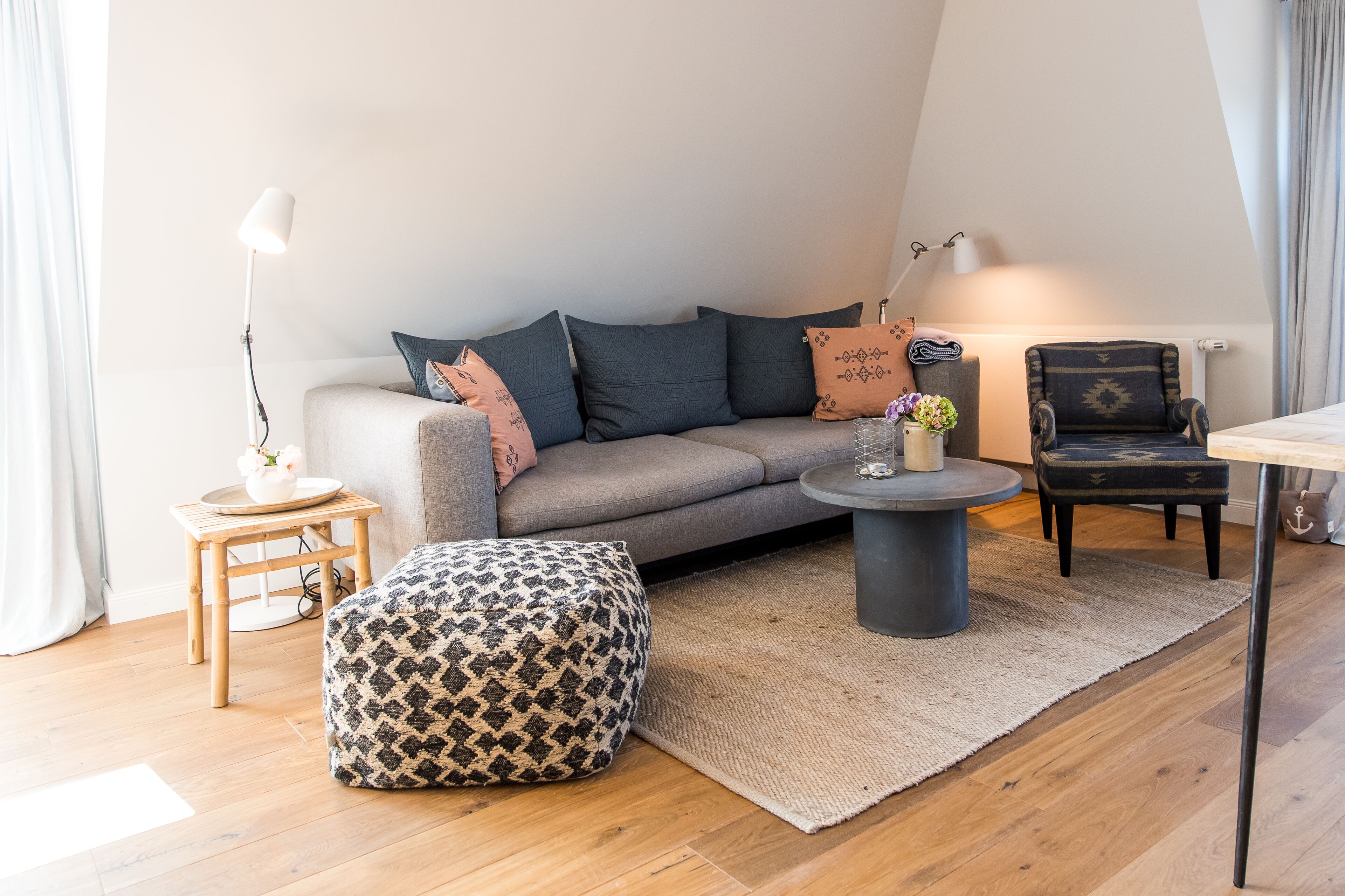 A modern living room with a grey sofa, patterned ottoman, round coffee table, and a small side table with a lamp.