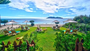 A beachside lounge area with lounge chairs and umbrellas, a grassy lawn, and a view of the sea and distant island. - Beach front "Andriana Villa" with mini pool and spa (Zakynthos)