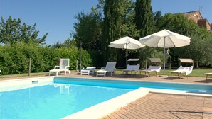 A swimming pool with lounge chairs and umbrellas, surrounded by trees and a building in the background. - La Dolce Collina (Castiglione del Lago)