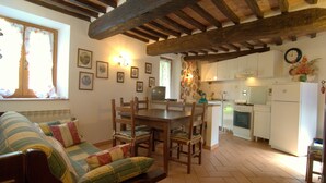 A cozy living room with a wooden dining table, chairs, and a sofa with patterned cushions. A kitchen with white appliances and a tiled floor is visible in the background. - La Dolce Collina (Castiglione del Lago)