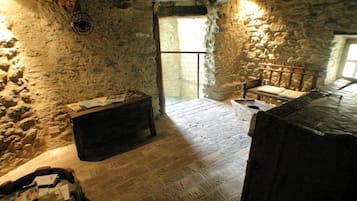 A stone-walled room with a wooden bench, a table with papers, and a window allowing natural light.