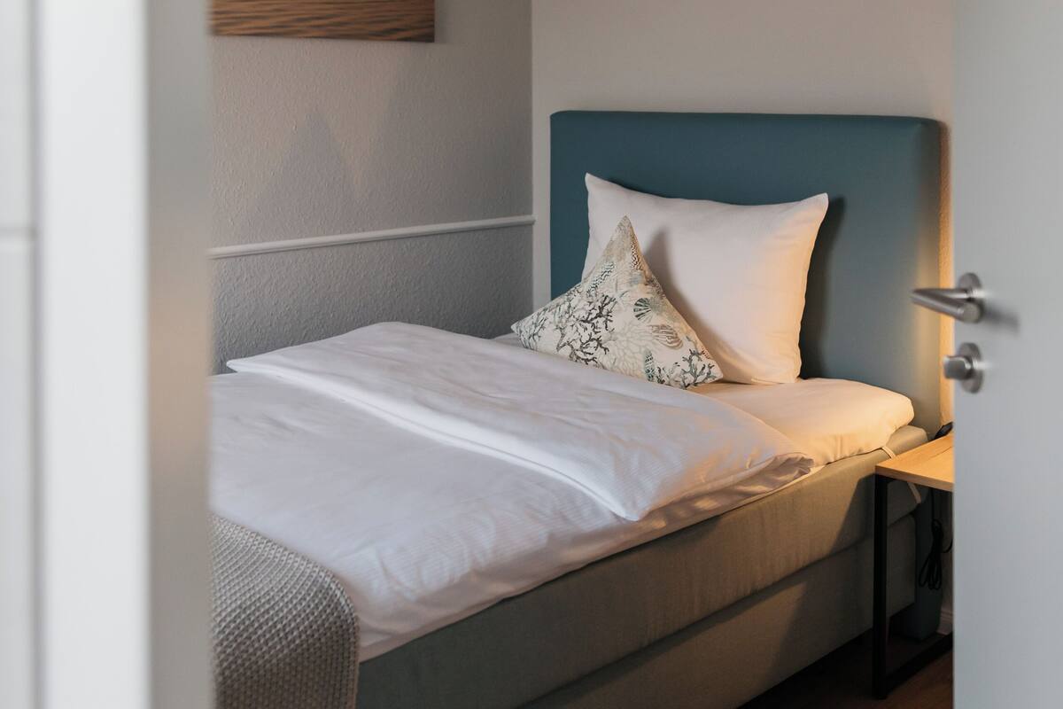 Un lit bien fait, avec du linge de lit blanc et un seul coussin décoratif, une table de chevet en bois avec une lampe, et un tableau représentant un paysage accroché au mur.