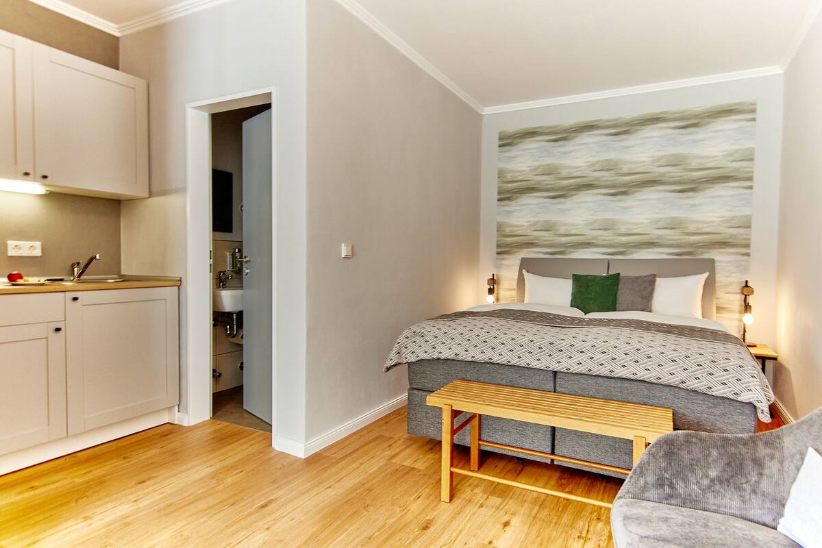 Une chambre à coucher de taille modeste, comprenant un lit, une table de chevet et donnant sur la cuisine.