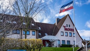 Een hotel met een groot 'HOTEL' bord, een vlaggenmast met een vlag, een trap naar de ingang en een bank op de voorgrond.