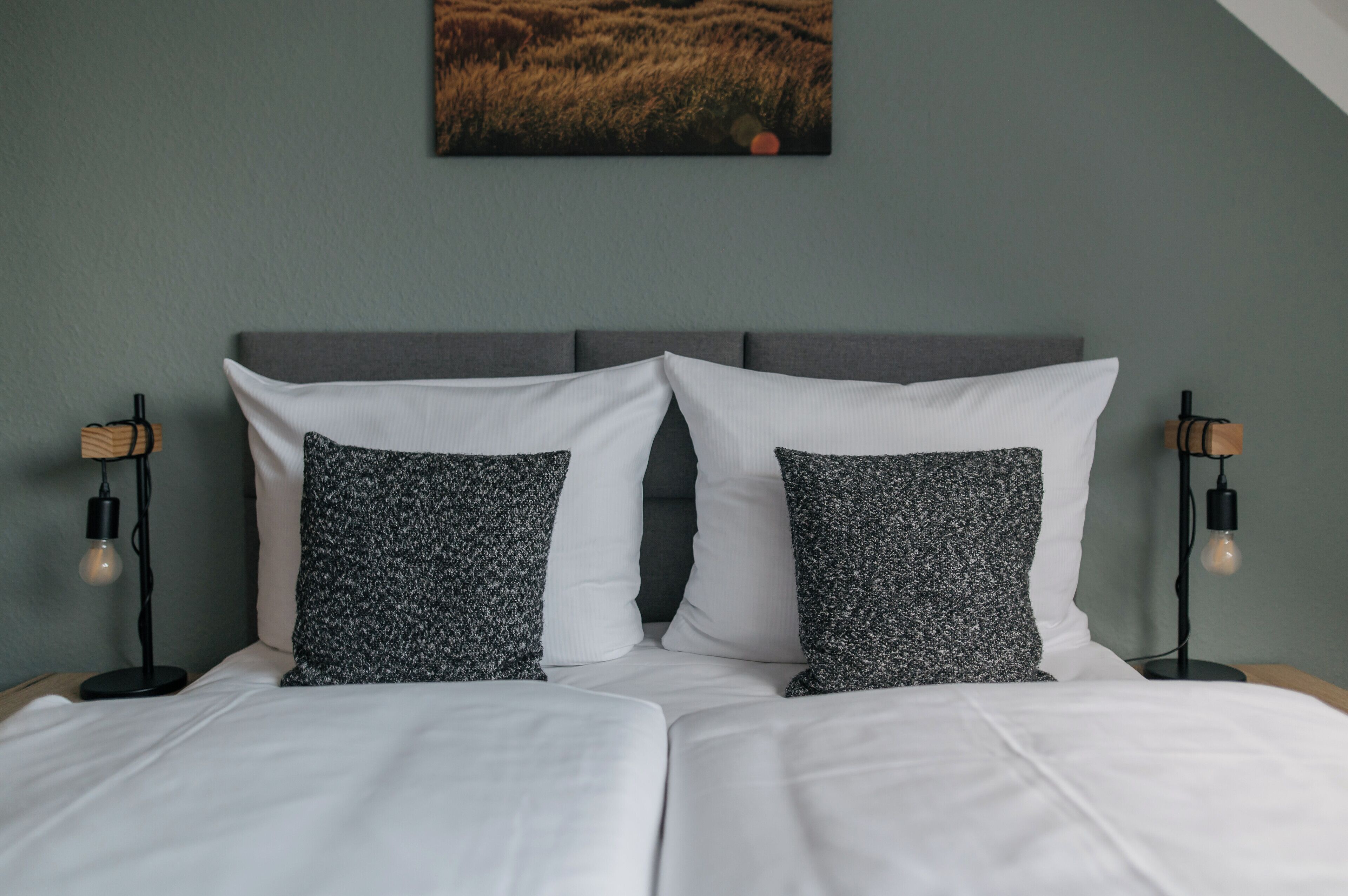 Una cama bien hecha con sábanas blancas y dos almohadas estampadas, con dos lámparas de noche a cada lado con bombillas expuestas.