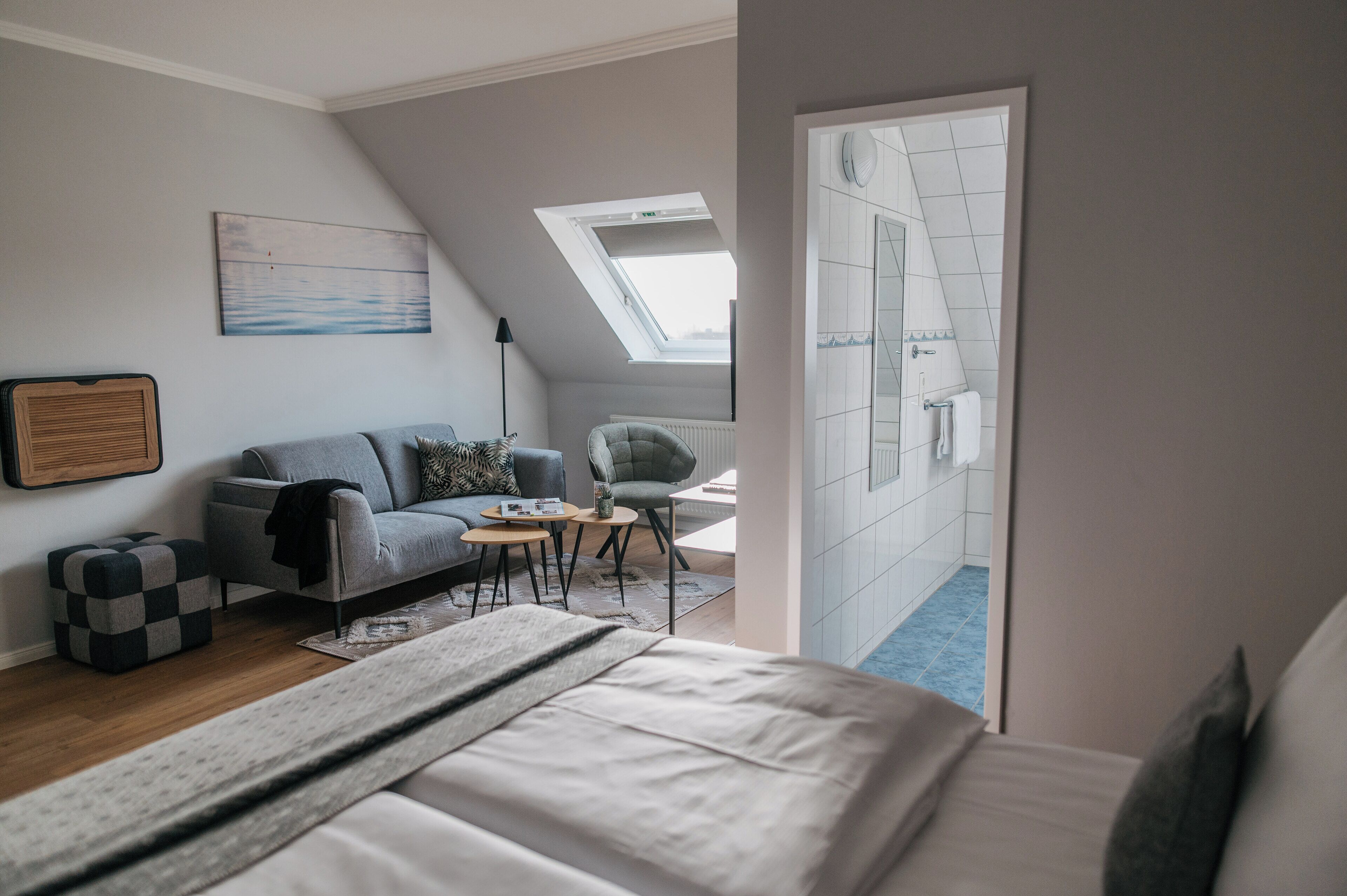 A modern bedroom with a bed, a gray sofa, a small table, and a chair. There is a skylight above the sofa and a view of the bathroom through an open door.