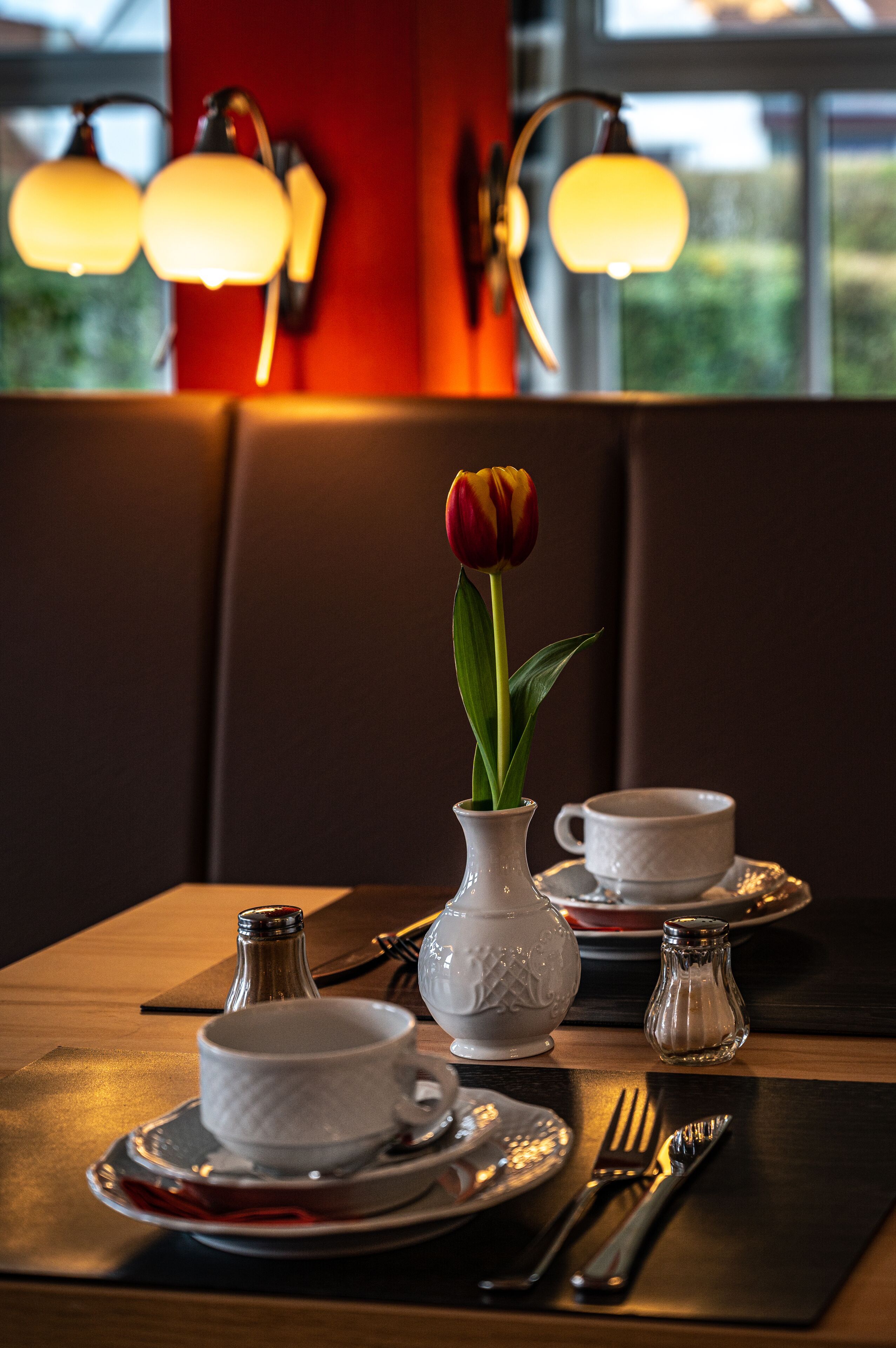 A dining table set with a white cup, saucer, and a small vase with a single tulip.