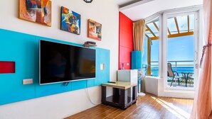 Un salon moderne doté d’un grand téléviseur à écran plat, d’œuvres d’art aux couleurs vives et offrant une vue sur la mer grâce à une porte-fenêtre coulissante.