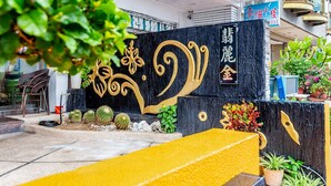 L’entrée d’un hôtel, avec un mur noir orné d’éléments décoratifs dorés et de caractères chinois. On aperçoit, au premier plan, des plantes en pot et un banc jaune.