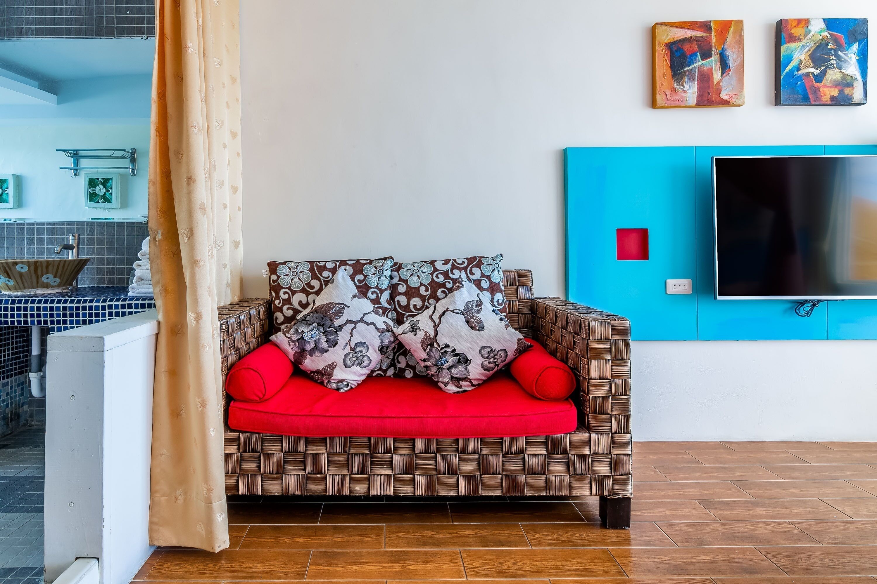 A living area with a woven sofa, red cushions, and a flat-screen TV mounted on a blue wall.