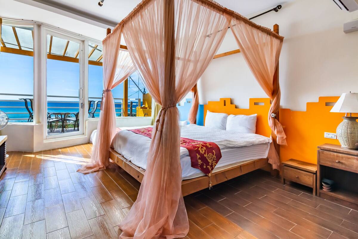 A bedroom with a four-poster bed, a wooden nightstand, and a view of the ocean through large windows.