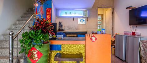 A hotel reception area with a staircase, a reception desk, a television, and a large green vase with a red seal.